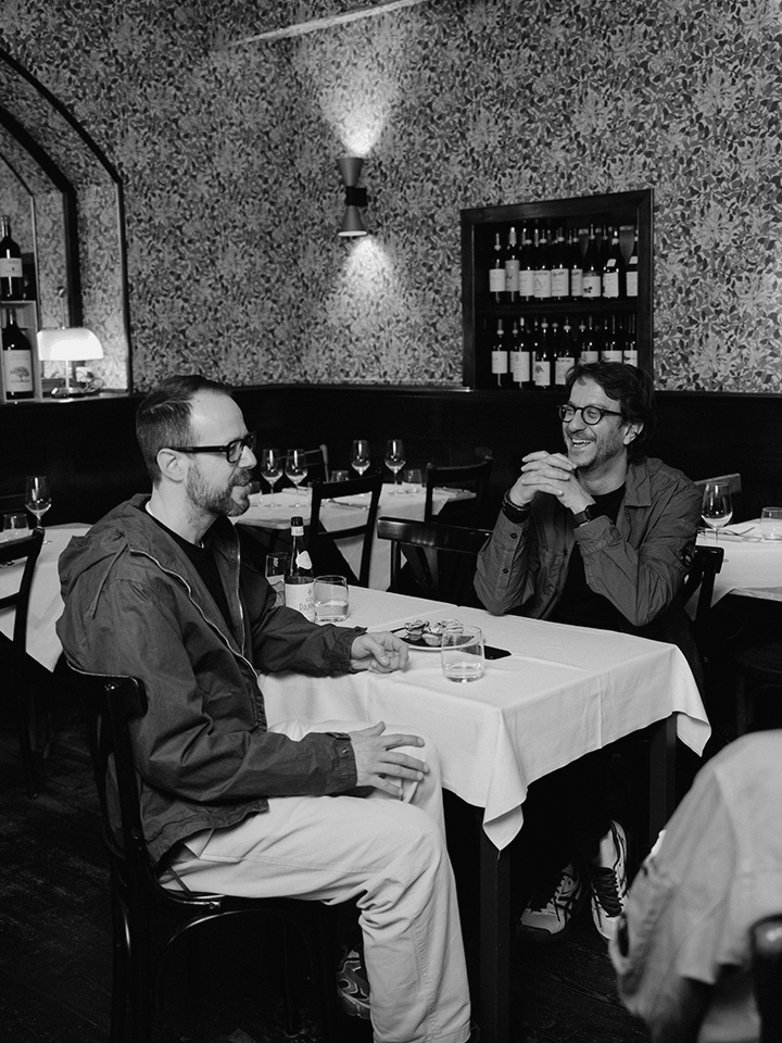 Two men in glasses laugh and converse at a cozy restaurant table, sharing ideas over drinks in warm, intimate lighting.