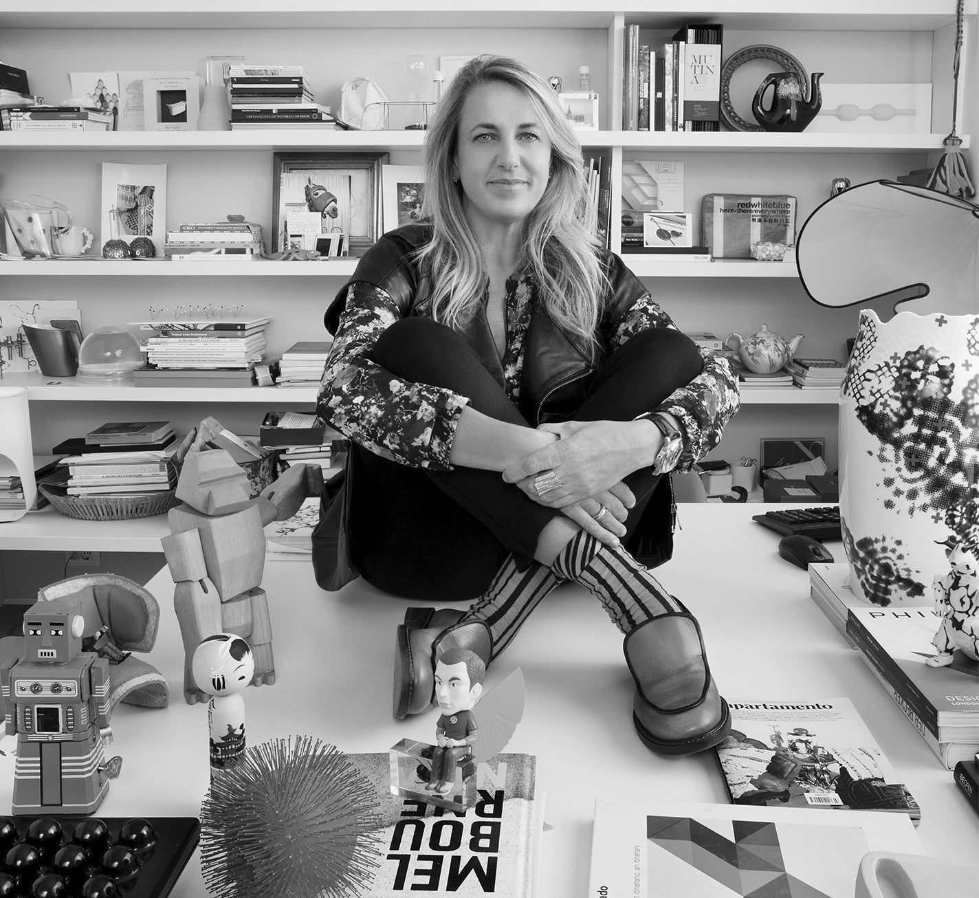 Patricia Urquiola sits casually on a desk surrounded by books, toys, and design objects in her creative studio.