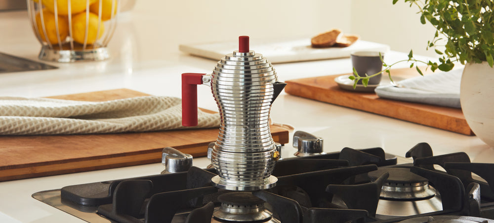 Shiny silver espresso maker with red handle on gas stove, surrounded by kitchen items and lemons.