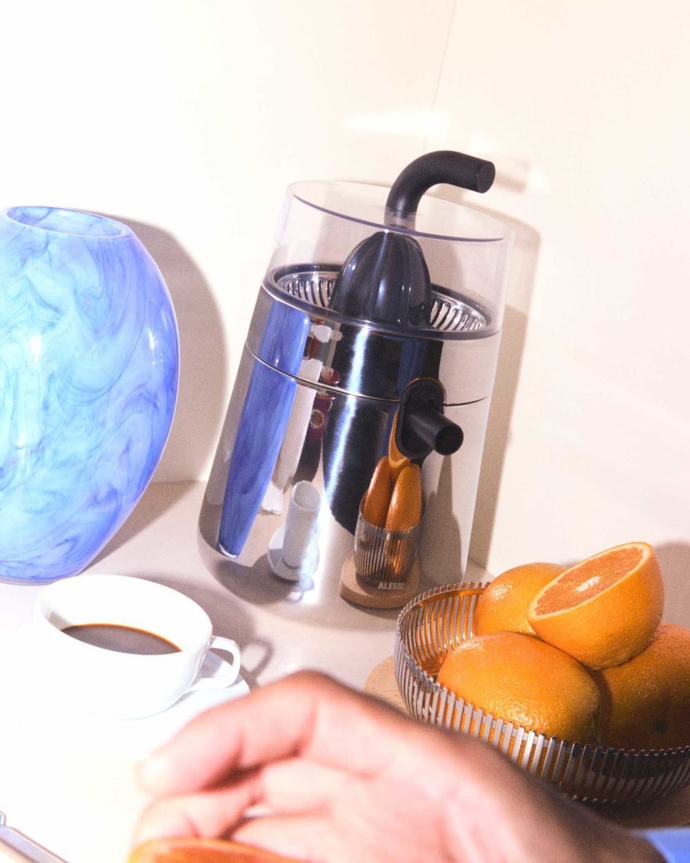Leaning silver and blue electric citrus squeezer by Alessi, featuring a black spout, surrounded by fresh oranges and a coffee cup.