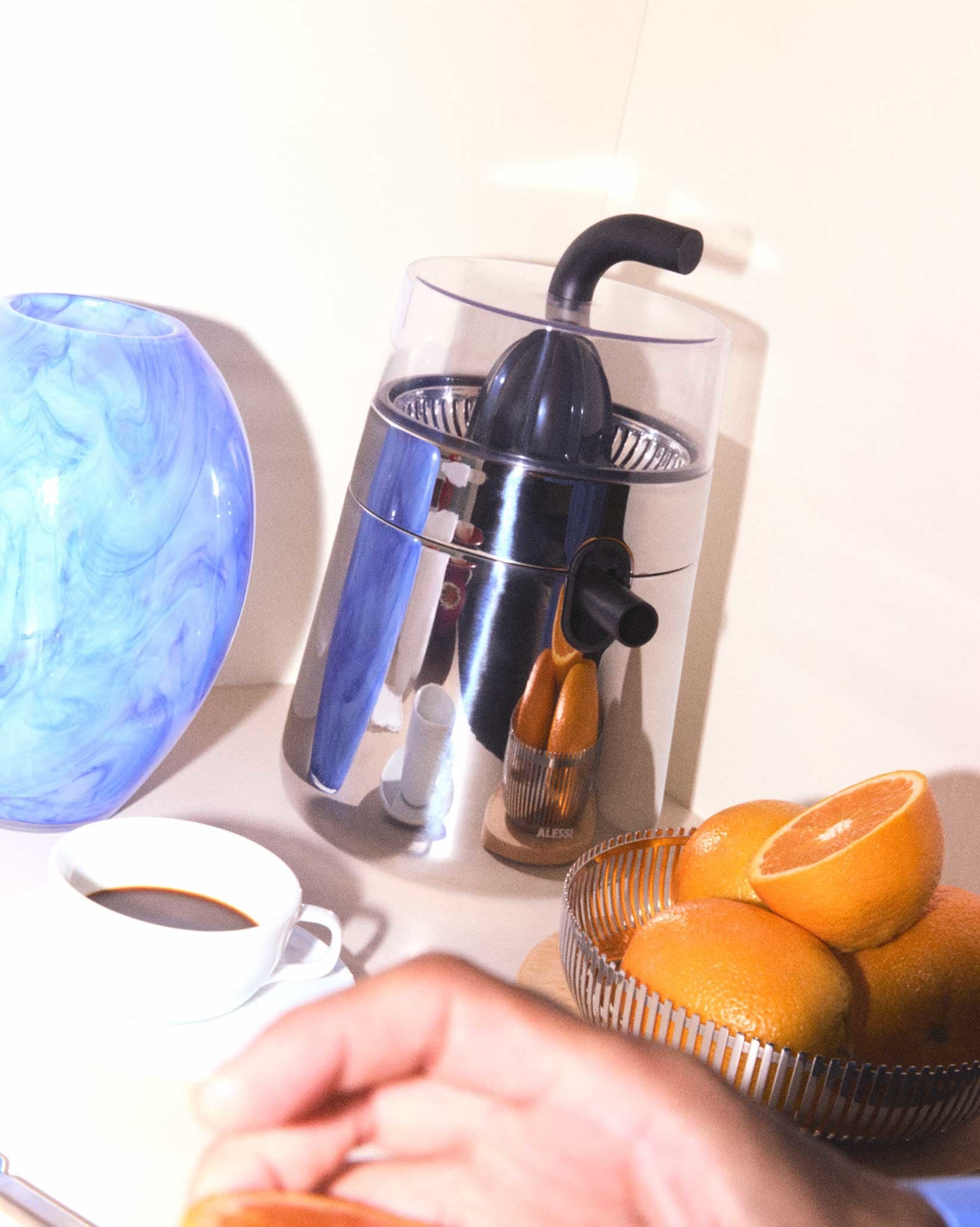 Leaning silver and blue electric citrus squeezer by Alessi, featuring a black spout, surrounded by fresh oranges and a coffee cup.