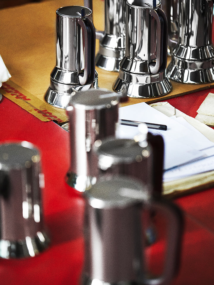 Shiny chrome coffee pots arranged on red tablecloth, with papers and pen nearby, warm lighting.