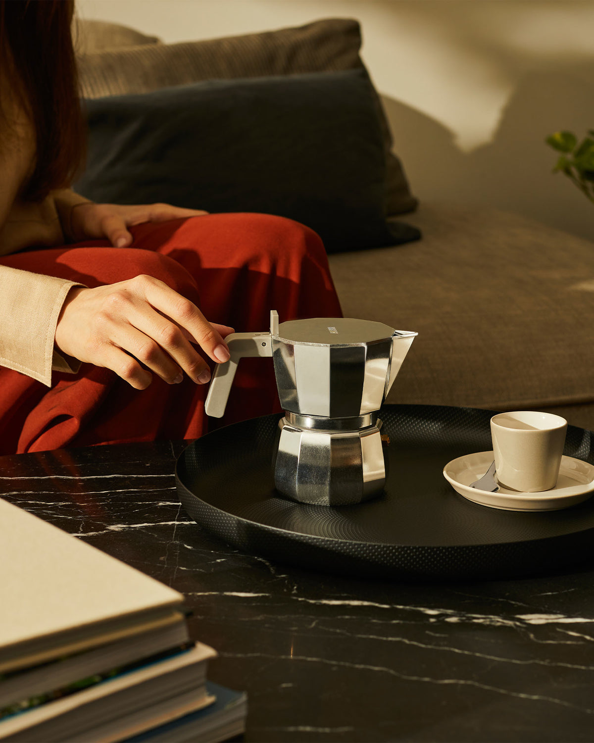 Silver octagonal Moka pot with a light gray handle by Alessi, held by a hand on a black textured tray with a coffee cup.